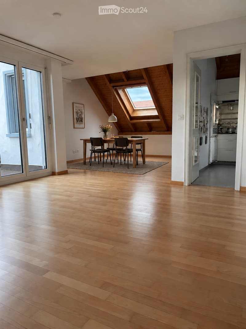 Bright, open-plan living and dining area with wooden flooring, large windows, skylight, and a modern kitchen in the background.
