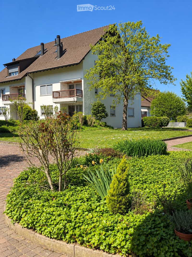 Two-story white house with a brown tiled roof, multiple windows, and balconies. The surrounding garden features well-maintained lawns, flowering plants, and a brick pathway.