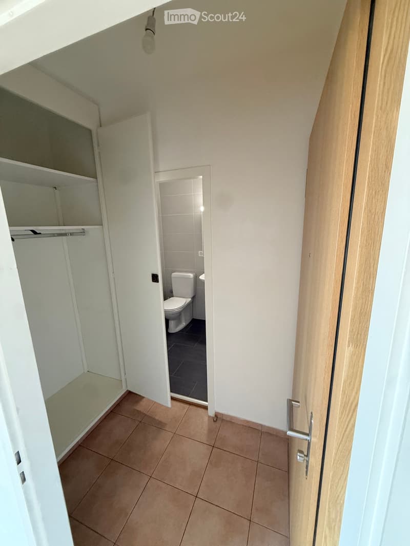 A narrow hallway with tiled flooring, white walls, and a wooden door leading to a small bathroom with a toilet and sink.
