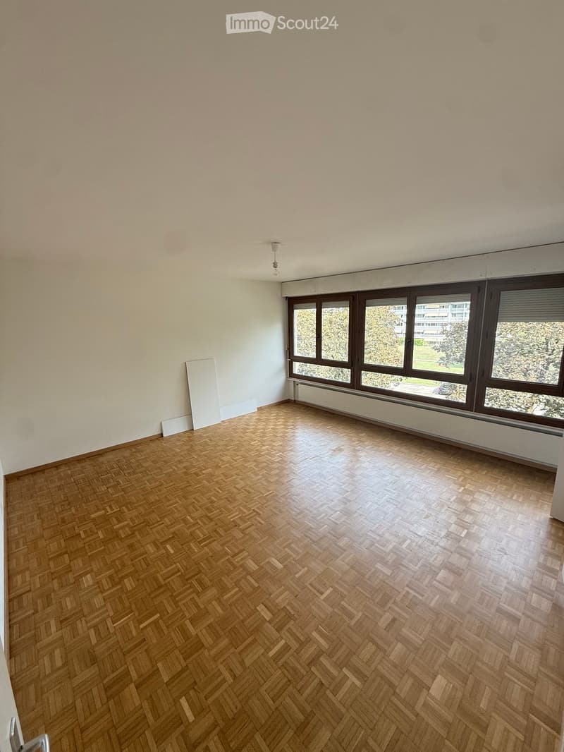 Empty room with wooden herringbone flooring, large windows with brown frames, and white walls.