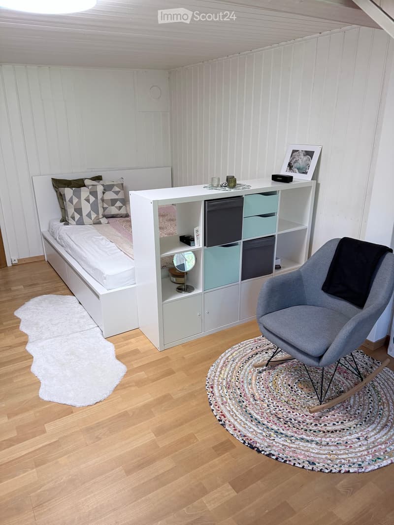 Cozy bedroom with a white bed, patterned pillows, a grey armchair, a white shelf unit with storage compartments, a round woven rug, and a white shaggy rug near the bed.