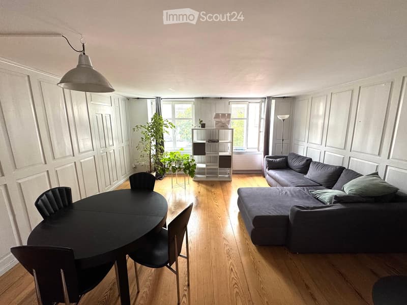 A modern, open-concept living and dining area with wooden flooring, large windows, a grey sectional sofa, a black dining table with chairs, and a pendant light fixture.