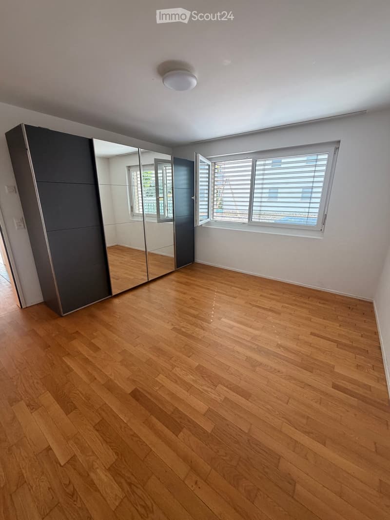Empty room with wooden flooring, white walls, large windows with blinds, and a mirrored wardrobe.