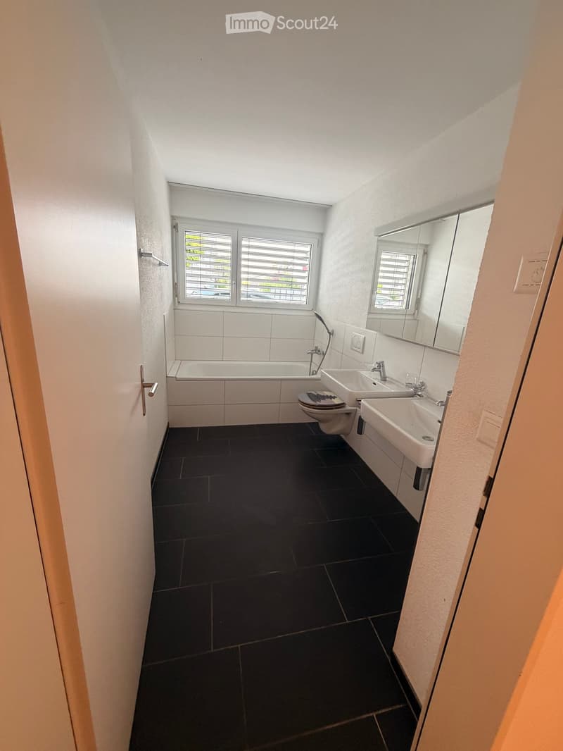 Modern bathroom with dark tiled floor, white tiled walls, bathtub, two sinks, toilet, and a large mirror above the sinks.