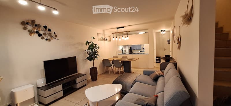 Modern open-plan living and dining area with a grey sectional sofa, coffee table, TV stand, dining table with chairs, and a small kitchen area visible in the background.