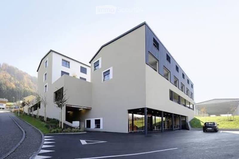 Modern multi-story building with clean lines, large windows, and a parking area in front.