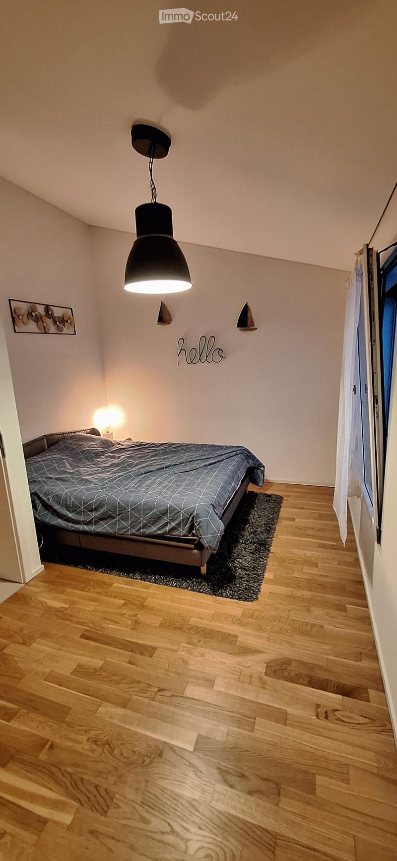 Cozy bedroom with wooden flooring, a bed with a grey duvet, a hanging pendant light, a small side table lamp, and a decorative rug.