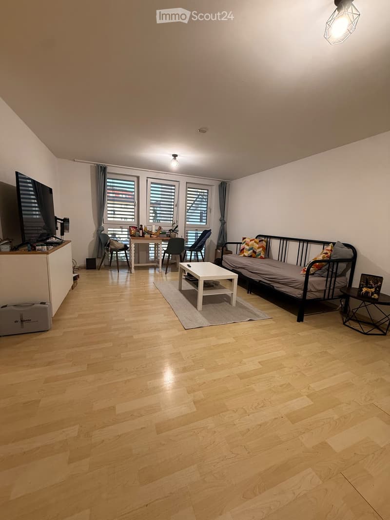 A modern, multi-functional room with a sofa bed, coffee table, small table, chairs, and a TV screen. The room features wooden flooring, large windows with blinds, and a cozy layout.