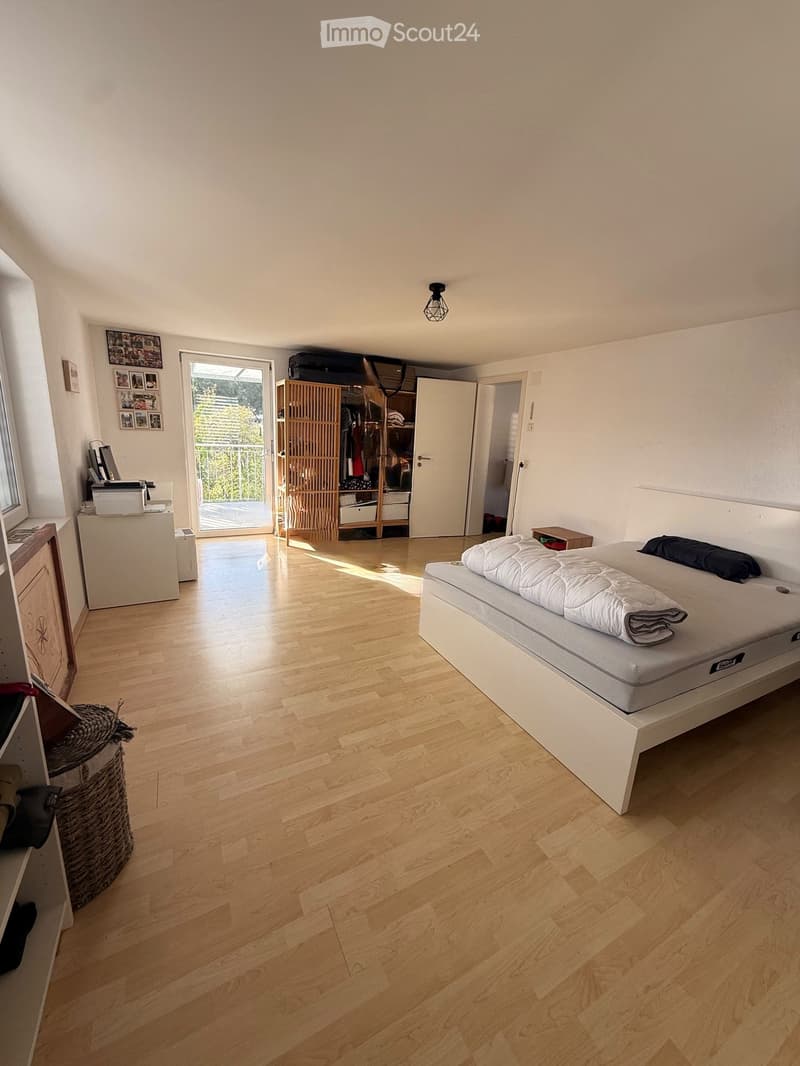 Bright bedroom with wooden flooring, a white bed with a quilt, a wicker basket, a wardrobe, and a printer on a small table. A sliding glass door leads to a balcony.