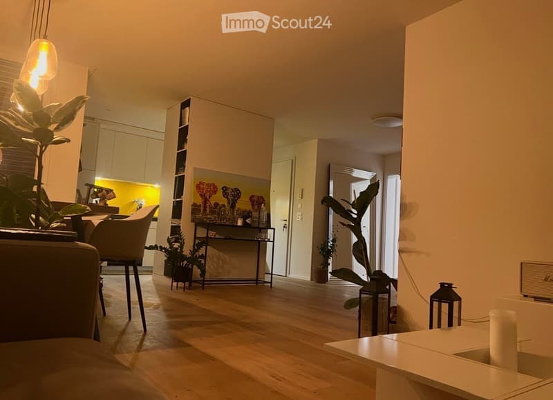 Modern living room with wooden flooring, white walls, and large windows. Features a sofa, chair, small table, shelf with decorative items, potted plants, and a picture frame on the wall.