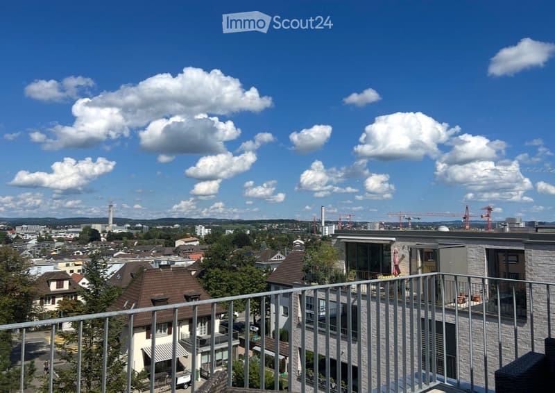 Balcony overlooking a cityscape with residential buildings, trees, and distant construction cranes under a partly cloudy sky.