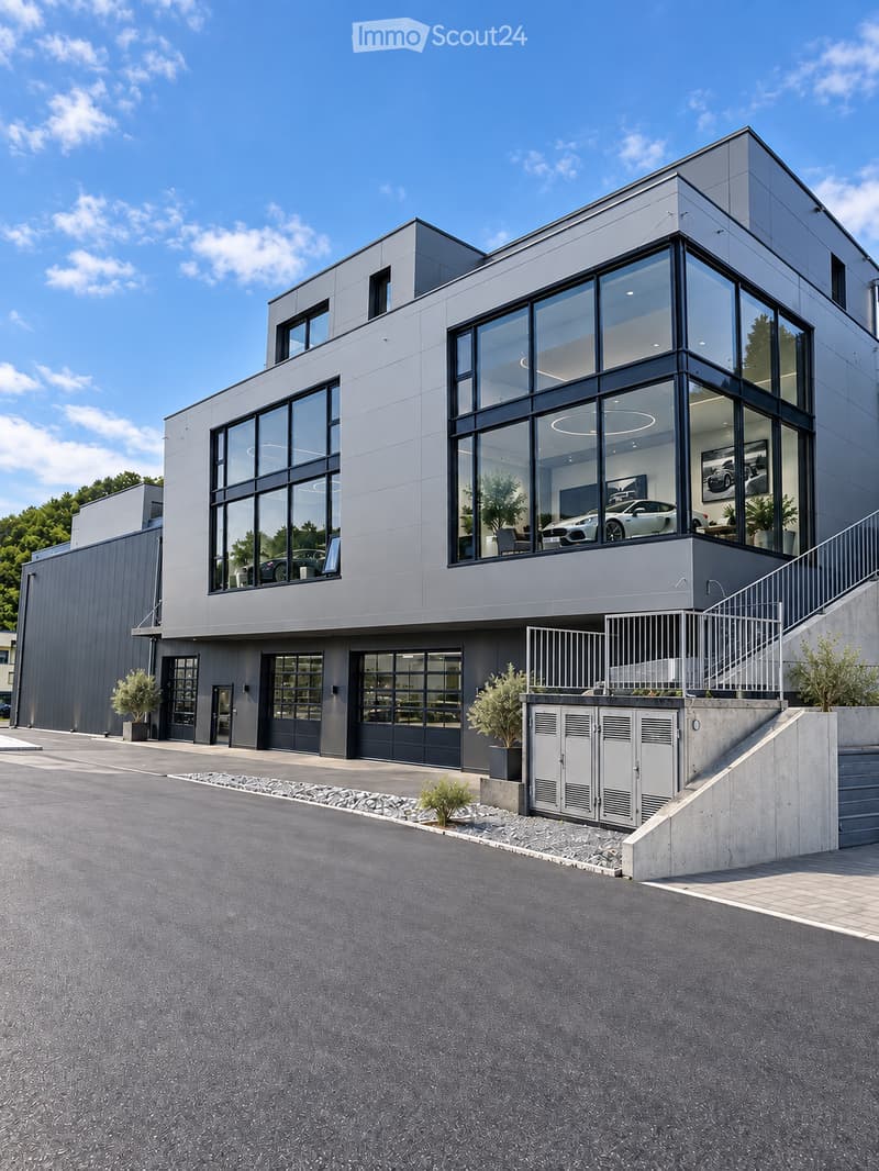 Modern two-story building with large glass windows and grey facade, featuring a raised ground floor, garage, and landscaped entrance area.