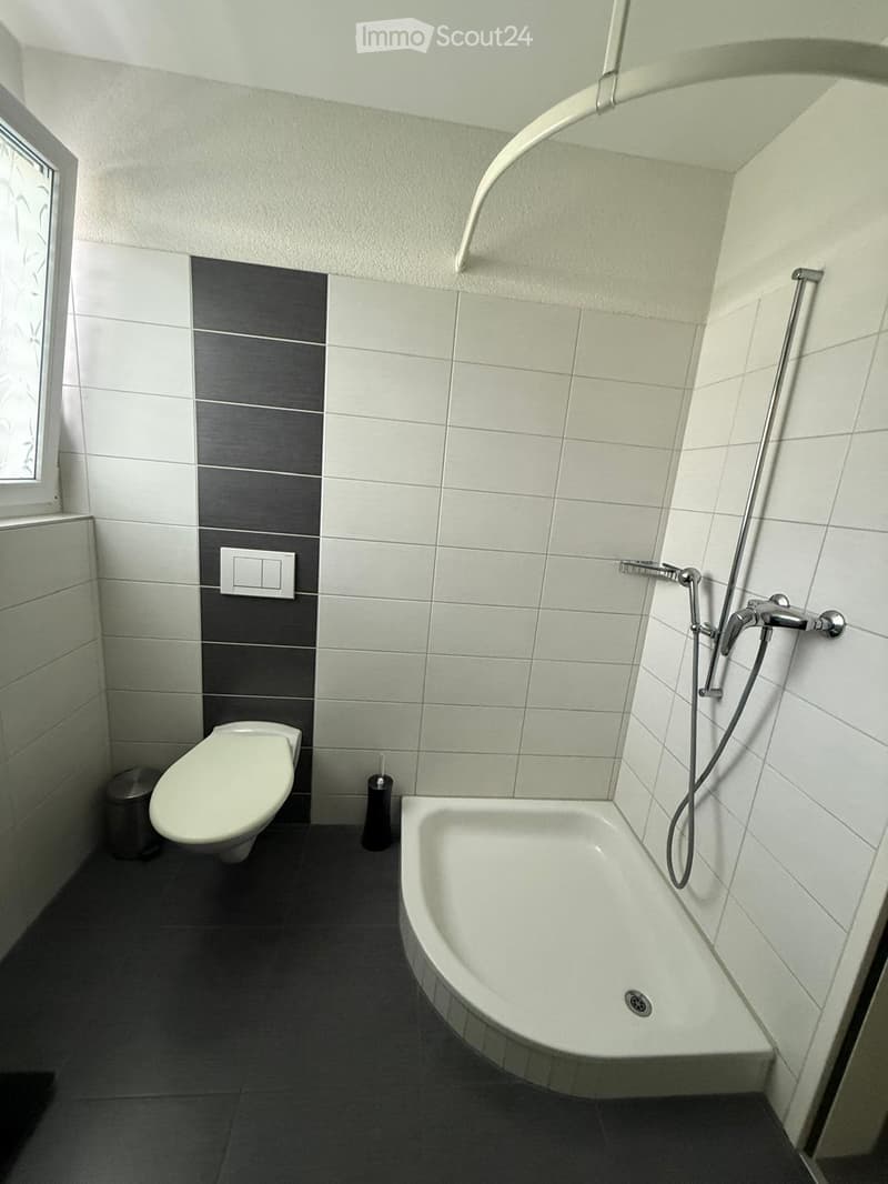 Modern bathroom with white tiles, dark grey accent wall, bathtub, toilet, shower hose, and a small trash can.