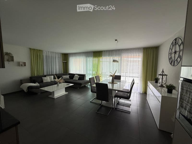 Modern living and dining area with dark grey sofa, white coffee table, glass dining table with chairs, large windows with curtains, and decorative items including a clock and sideboard.