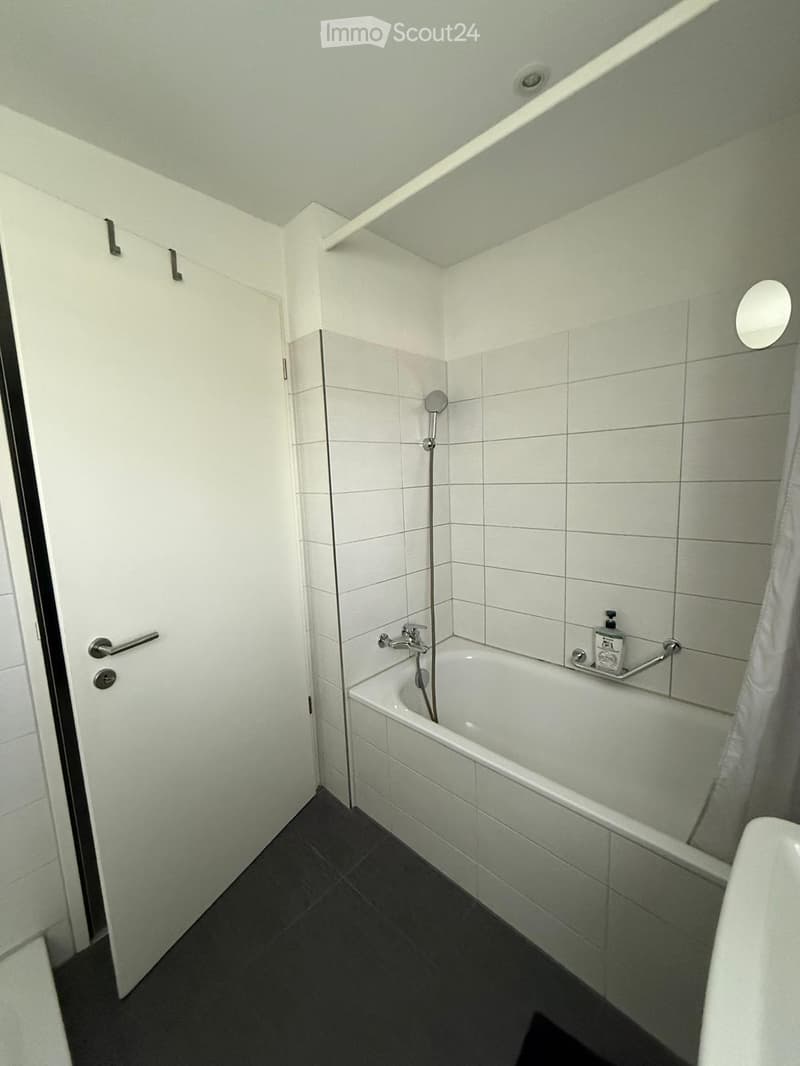 Modern bathroom with white tiled walls, a bathtub, a showerhead, a small mirror, and a bottle on the edge of the tub.