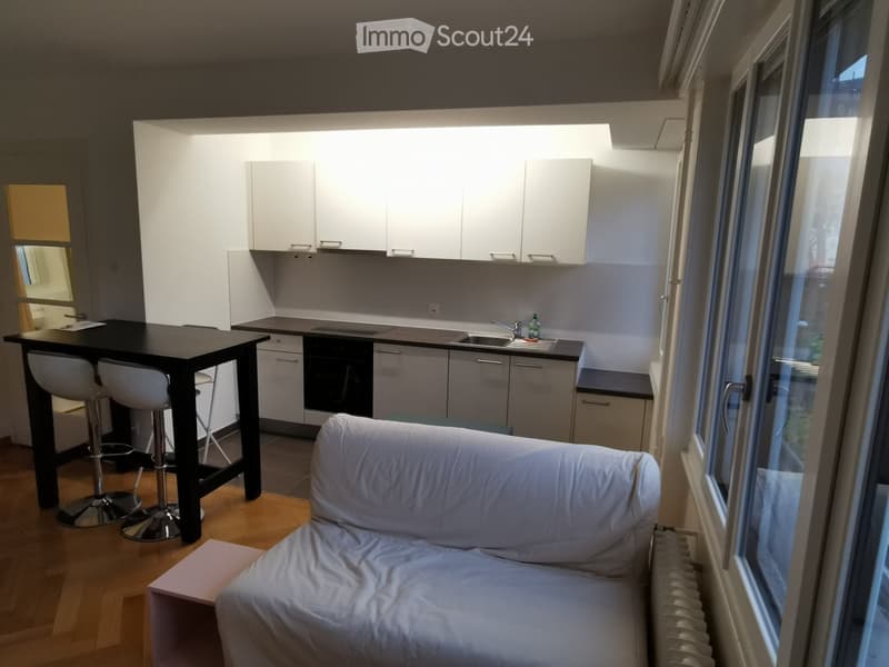 A modern, open-plan living and dining area with a white sofa, a black dining table with four white stool chairs, and a sleek kitchen featuring white cabinets, a sink, and a faucet.