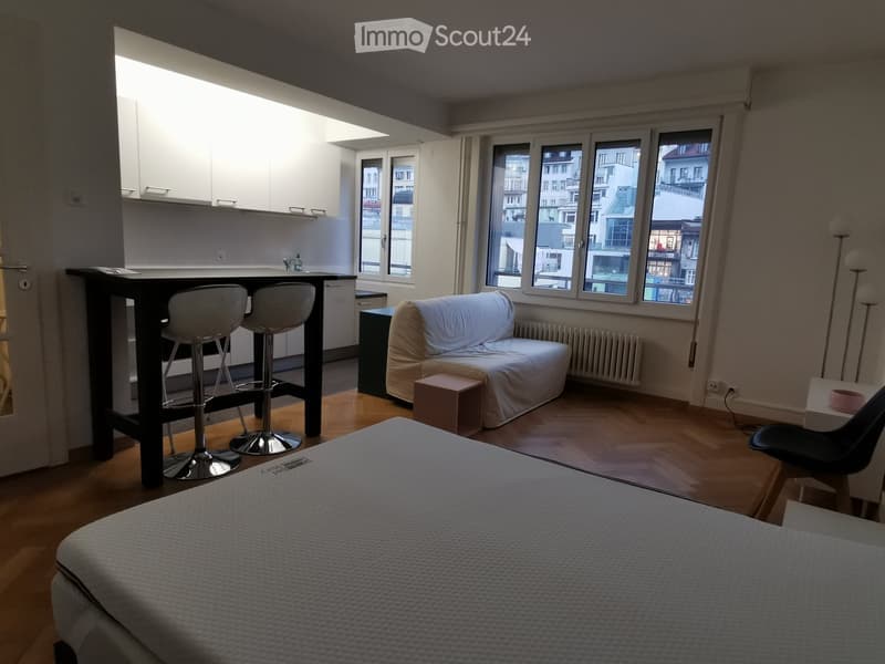 A modern, open-concept living space with a bed, sofa, and kitchen area. The room features large windows with city views, wooden flooring, and contemporary furniture including a sofa, a small table, and bar stools.