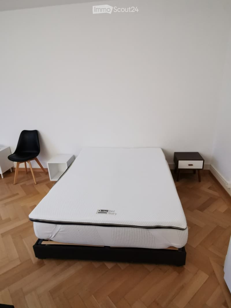 A furnished bedroom with a white mattress on a black base, a black chair, and a small nightstand.