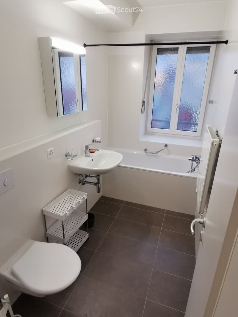 Modern bathroom with white walls, dark tiled floor, sink, bathtub, toilet, mirror, and a small white shelf.