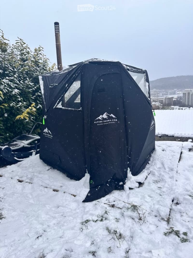 A black tent with a chimney is set up in a snowy outdoor area, surrounded by snow-covered ground and bushes.