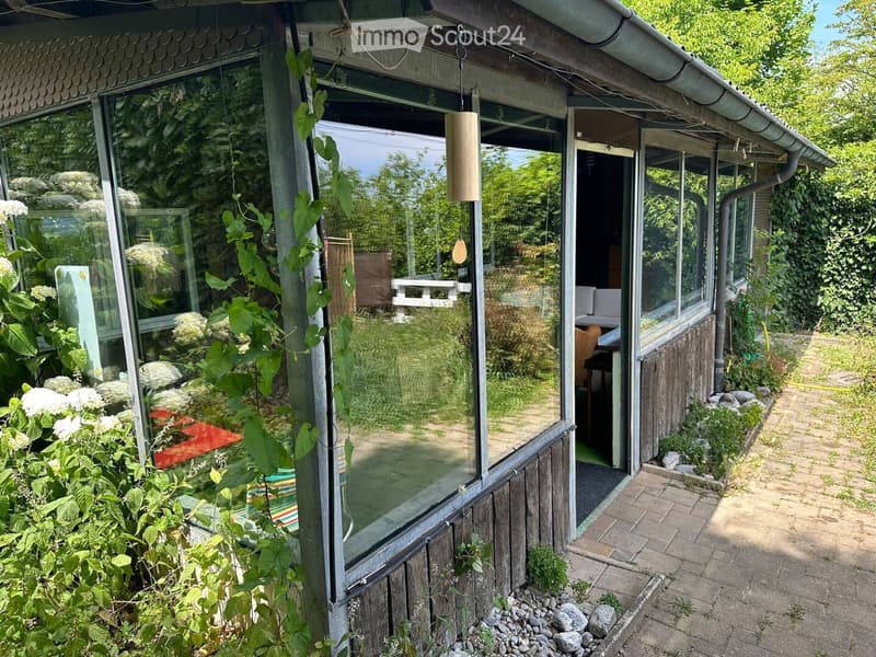 A glass-enclosed patio area with wooden frame, surrounded by lush greenery and flowers, with a hanging wooden ornament and a view of the garden.