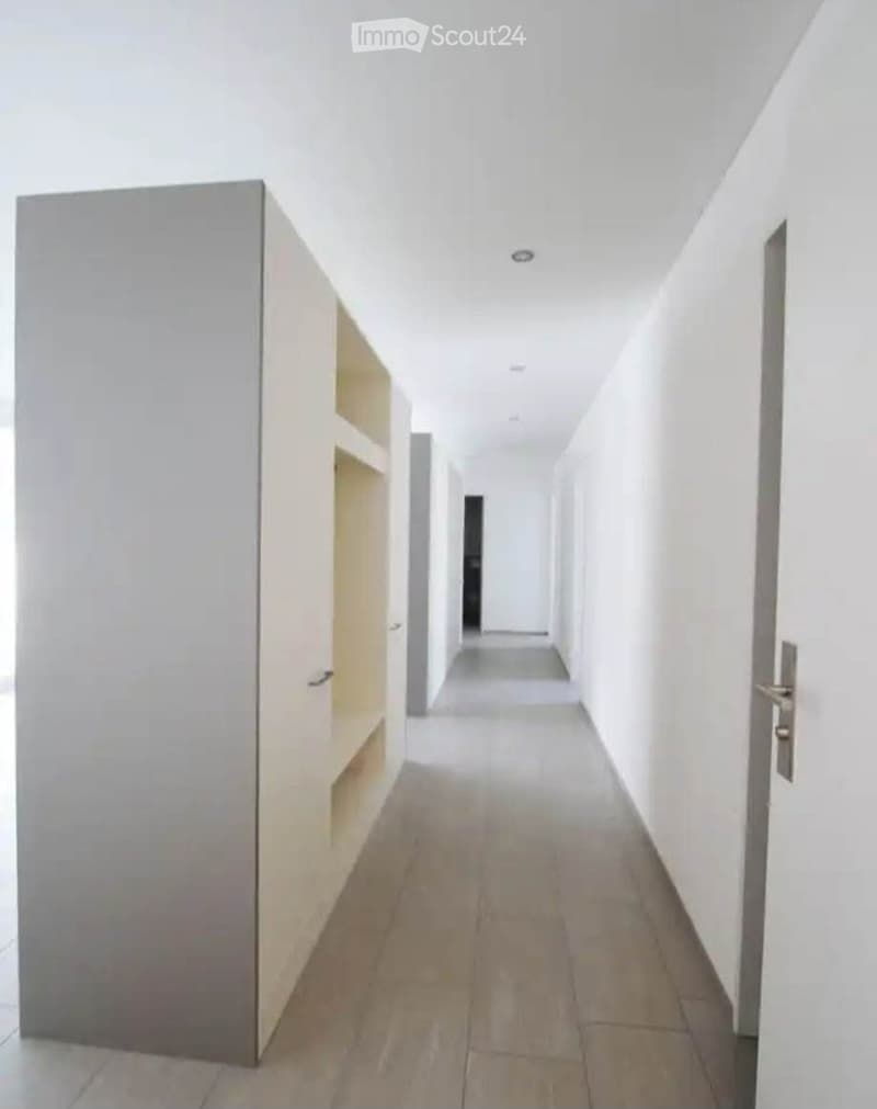 A clean, modern hallway with white walls, tiled flooring, and built-in shelving on one side. The corridor leads to a doorway at the far end.