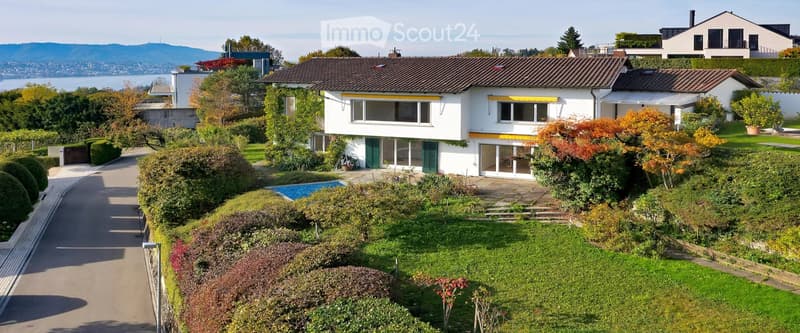 Aerial view of a two-story white house with a tiled roof, surrounded by a well-maintained garden, a swimming pool, and a scenic view of a lake and mountains in the background.