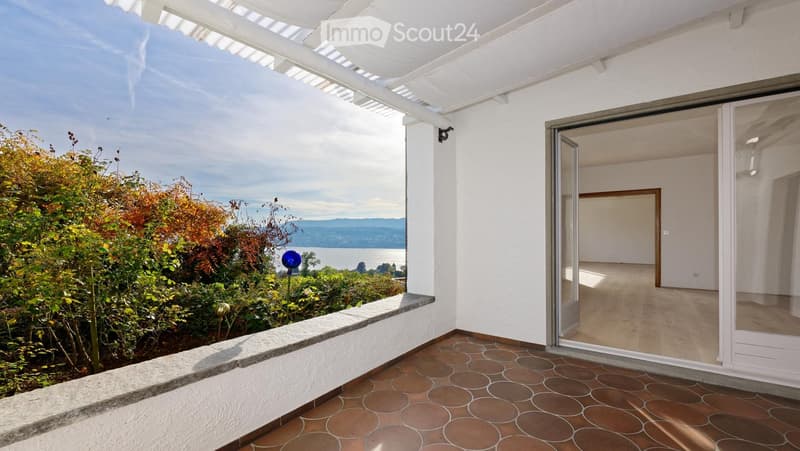 Balcony with tiled floor, white walls, open sliding glass door leading to an unfurnished room, scenic view of water and hills.