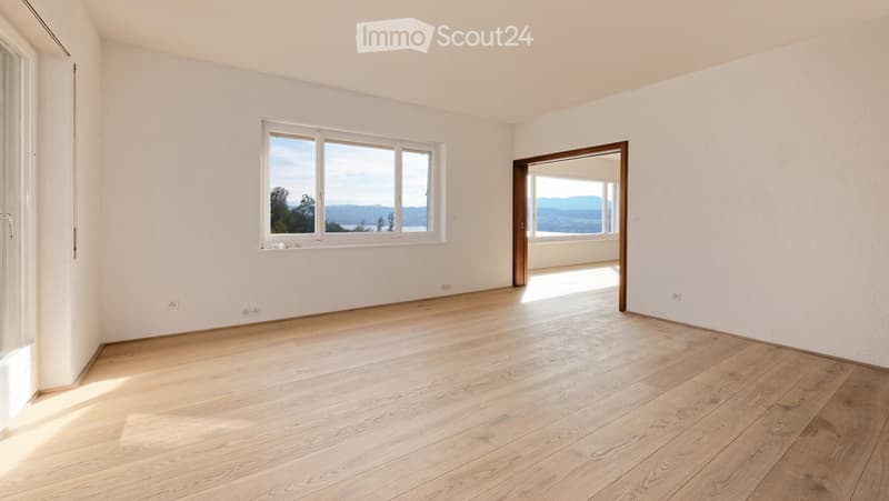 Empty room with light wooden flooring, white walls, and large windows offering scenic views.