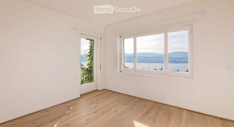 Empty room with white walls, wooden floor, large windows offering a scenic view of a lake and mountains.