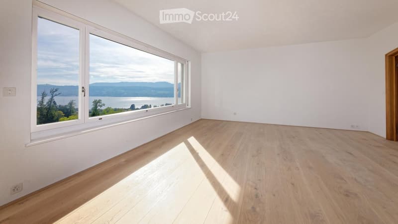 Empty room with light wooden flooring and large windows offering a scenic view of a lake and mountains.