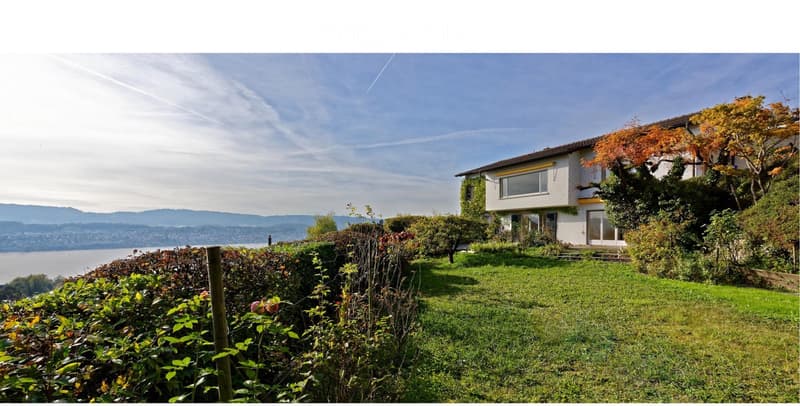 A two-story house with white walls and large windows, surrounded by a well-maintained garden and trees, overlooking a scenic lake and distant mountains.