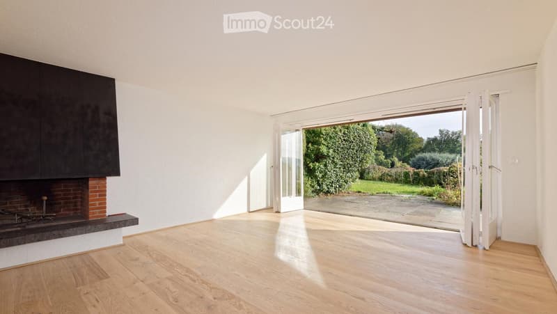 Empty room with wooden flooring, white walls, and a brick fireplace. Large sliding glass doors open to a garden.