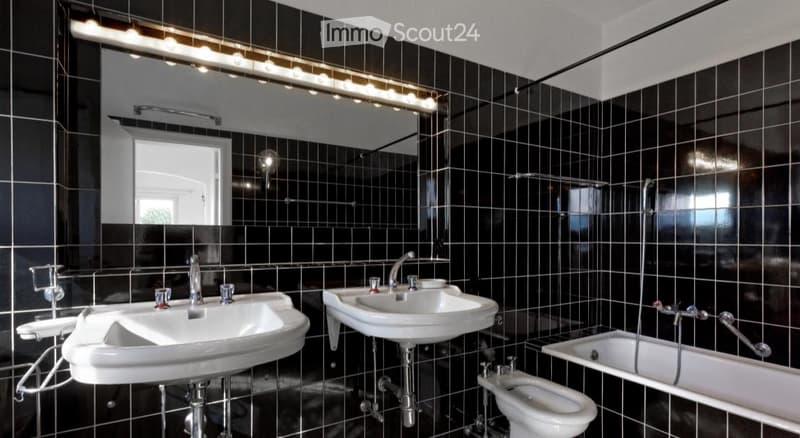 Modern bathroom with black tiled walls, two white sinks, a bathtub, and a toilet. Features include a large mirror with integrated lighting and a sleek faucet design.