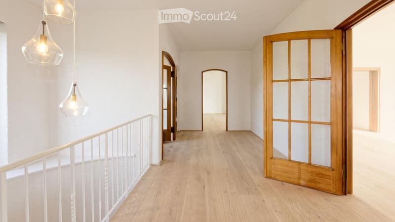 Bright, modern hallway with light wooden flooring, white walls, and pendant lights. Features include a white railing on the left and several doorways leading to other rooms.