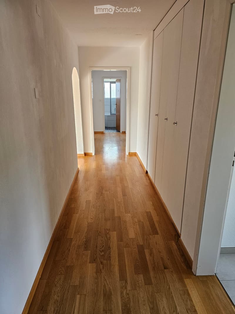A long, narrow hallway with wooden flooring and white walls, featuring several doors and a visible room at the end.