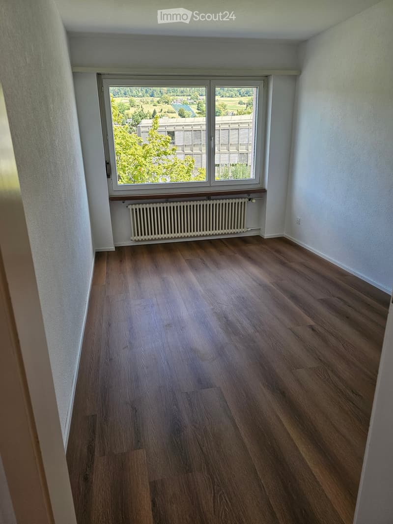 Empty room with wooden flooring, white walls, a radiator, and a large window offering a view of greenery and a building.