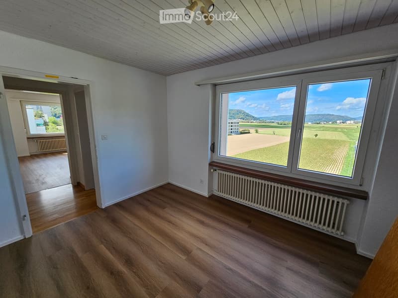 Empty room with wooden flooring, white walls, large windows offering a scenic view, and a radiator beneath the windows.