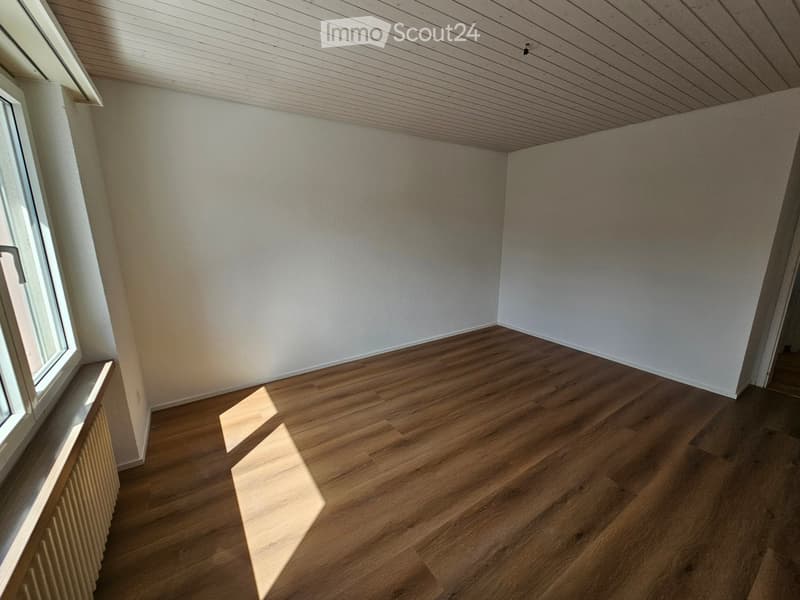 Empty room with wooden flooring, white walls, and a window on the left side. The room has a slanted wooden ceiling and a radiator below the window.