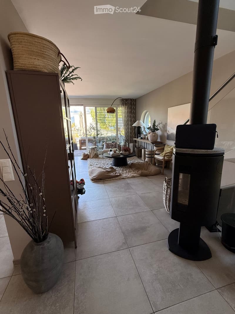 Modern living room with large windows and a balcony, featuring a fireplace, a round coffee table, chairs, a large vase with dried branches, and a mirror on the wall.