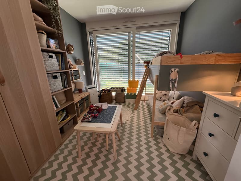 A well-lit children's bedroom with a bunk bed, a small table, a chair, shelves, and various toys and storage items.
