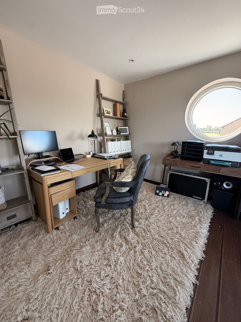 A well-furnished home office with a large wooden desk, ergonomic chair, laptop, monitor, printer, and shelves holding books and decorative items. The room features a plush white shag rug and warm lighting.