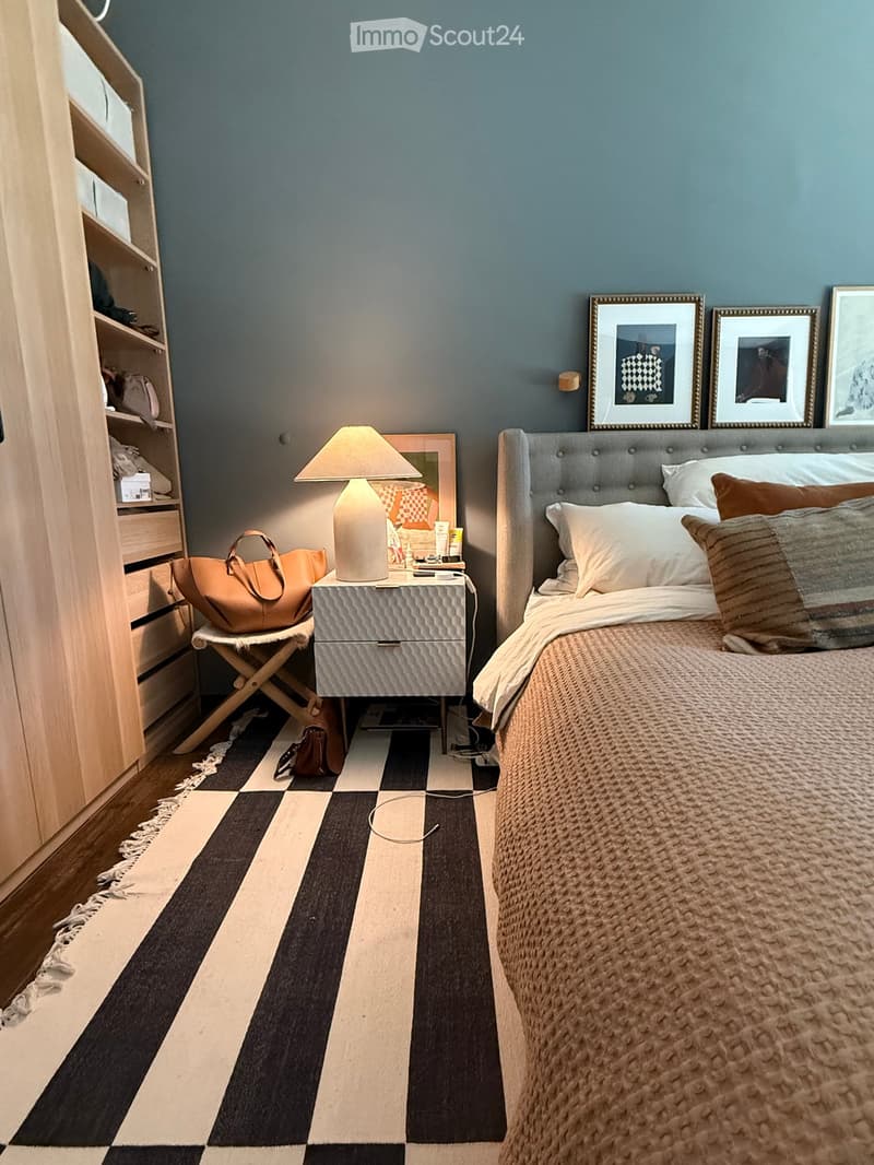 Modern bedroom with a tufted headboard bed, nightstand with lamp, mirror, and framed pictures on the wall. A wooden shelf unit is visible on the left side.