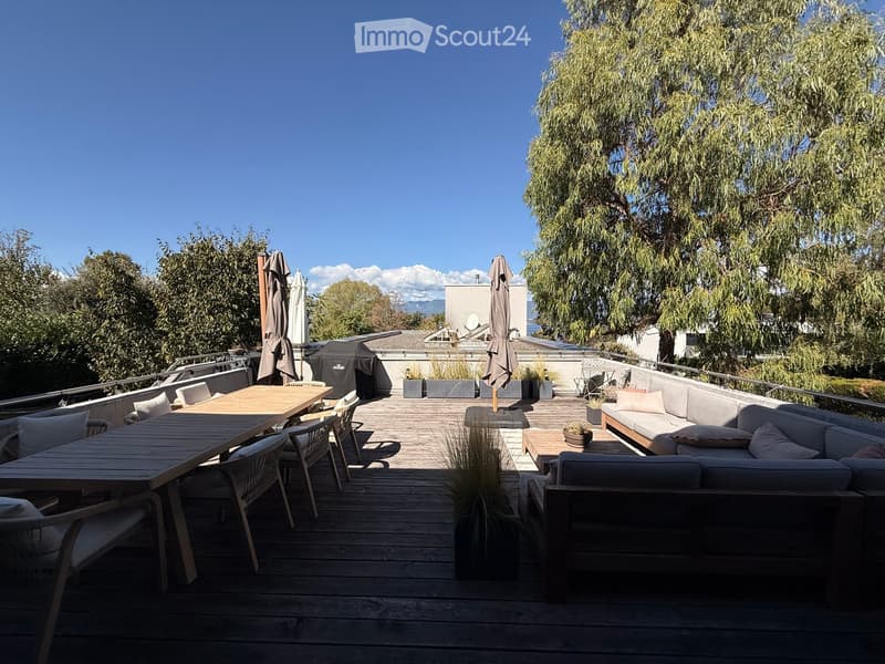 A spacious wooden deck with a sofa, dining table, chairs, and a grill, surrounded by trees and a clear blue sky.