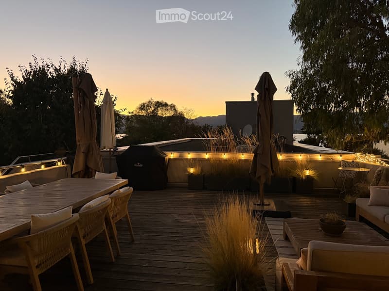 A wooden deck terrace with outdoor furniture, including a dining table, chairs, and a sofa, surrounded by potted plants and string lights, with a scenic sunset view in the background.