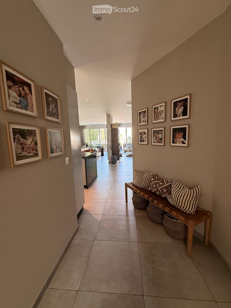 Hallway with beige tiled floor, wooden bench with striped cushions, framed family photos on walls, and a view into a living area with furniture and plants.