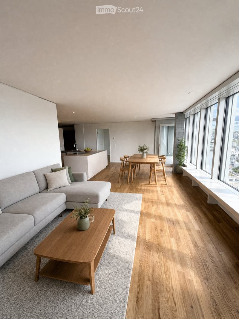 Modern living and dining area with light wood flooring, grey sectional sofa, wooden coffee table, dining table with chairs, and a small kitchen area visible in the background.