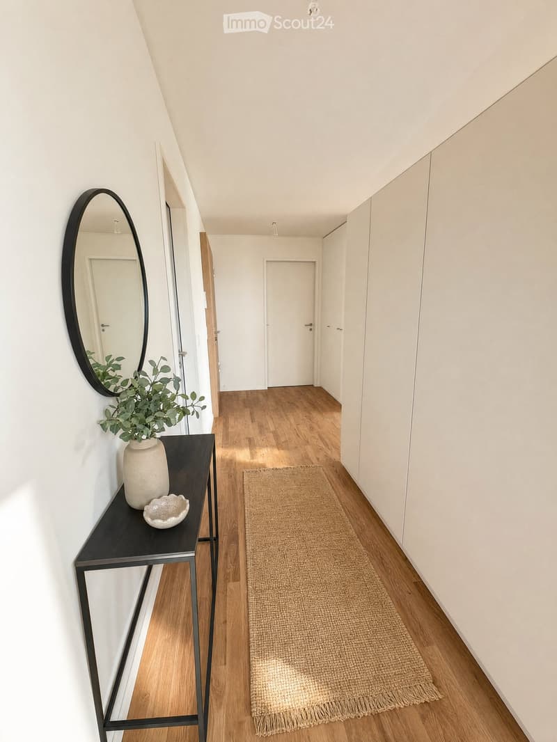 A modern hallway with wooden flooring, white walls, built-in wardrobes, a small black console table with a vase and bowl, a round mirror, and a natural fiber runner rug.