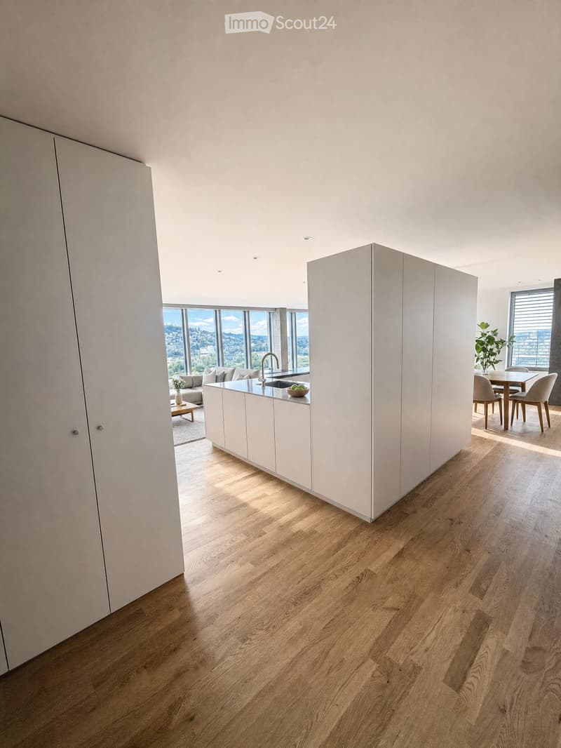 Open-plan living and dining area with modern white kitchen cabinets, wooden flooring, large windows offering a scenic view, and contemporary furniture including a sofa, coffee table, dining table, and chairs.