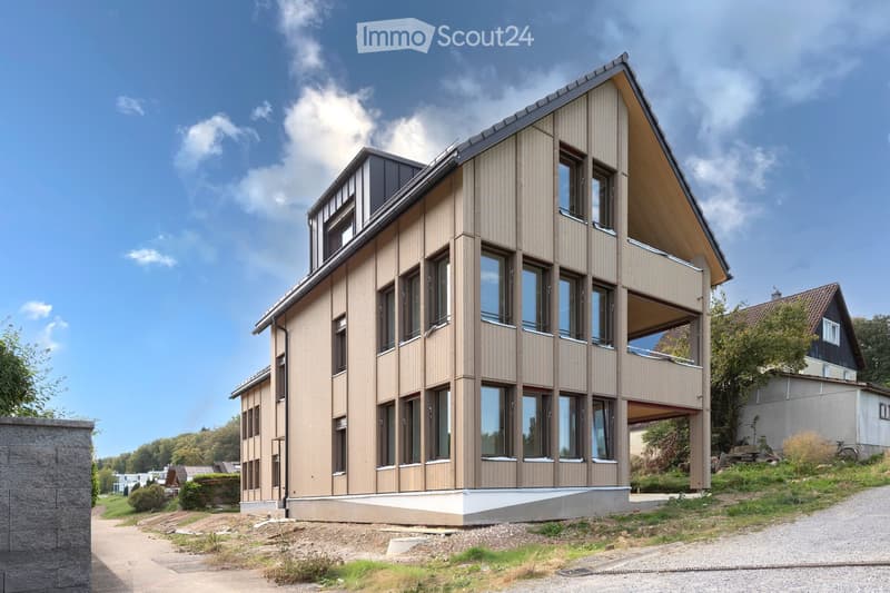 Modern two-story house with light brown wooden siding, large windows, and a raised ground floor. The house features a prominent balcony on the upper level and a sloped roof with dark accents.
